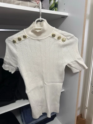Vit ribbad polotopp med knappar - Säljer en vit ribbad polotopp med korta ärmar och dekorativa guldknappar på axlarna. Toppen har en hög krage och är figurnära i modellen. Perfekt att styla med jeans eller kjol för en clean och trendig look.