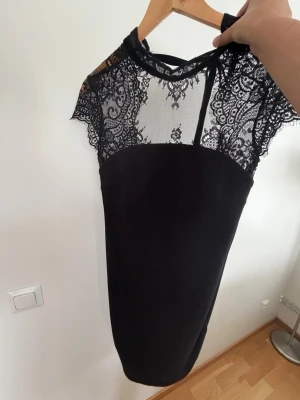 Svart klänning med spetsdetaljer - Elegant svart klänning med genombruten spets upptill och korta ärmar. Klänningen har en åtsittande passform och en klassisk, stilren look med vackra spetsmönster över axlar och bröst. Perfekt för dig som vill ha en chic och tidlös outfit.