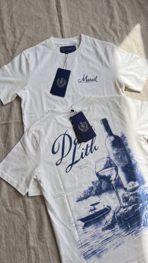 Vit t-shirt från Leon Marcel - Vit t-shirt från Leon Marcel med blått tryck. Framsidan har diskret logga och baksidan har ett stort motiv med vinflaska, glas och båt i blått. Klassisk rund hals och korta ärmar. Perfekt för dig som gillar stilrena men ändå unika plagg.
