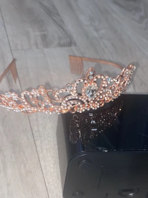 Diadem i roséguld med strassdetaljer - Säljer ett elegant diadem i roséguld med gnistrande strass och pärldetaljer. Diademet har en klassisk tiaraform med vackra utskurna mönster och stora stenar i mitten. även olika smycken.