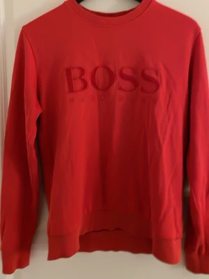 Röd sweatshirt från BOSS - Snygg röd sweatshirt från BOSS med broderad logga framtill. Tröjan har rund halsringning, långa ärmar och ribbade muddar vid ärmslut och nederkant. Perfekt för en avslappnad och trendig stil.