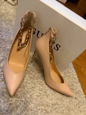 Nude pumps med guldkedja från Guess - Säljer ett par nude pumps från Guess med spetsig tå och hög smal klack. Skorna har en chunky guldfärgad kedja runt ankeln som ger en lyxig touch. Köpta för 1870kr eller 170€. Lätt använda en gång i någon timme (beige rosa färg ) 