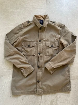 Beige overshirt från Gap - Stilren khaki/oliv grön overshirt från Gap med två bröstfickor med lock och knappar, axelklaffar och klassisk krage. Storlek M, tveka inte att skriva vid funderingar!!