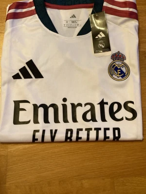 Real Madrid matchtröja 2026-2027 Adidas vit - Snygg vit Real Madrid matchtröja från Adidas med klubbmärke och sponsortryck på bröstet. Tröjan har svarta detaljer, mörkgröna muddar och vinröda ränder på axlarna. Tillverkad i lätt och ventilerande material, perfekt för fotbollsträning eller match.
