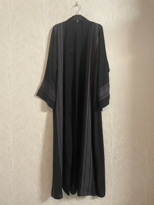 Svart Abaya från Saudiarabien - En svart abaya köpt från Saudiarabien som har använts ett par gånger men ser fortfarande ut som ny! Den har vida ärmar och diskreta grå ränder med glittriga strassdetaljer (några har ramlat av). Klänningen har en rak/vid och lös passform med knäppning framtill. Längd: ca 130 cm. Bredd: ca 60 cm.