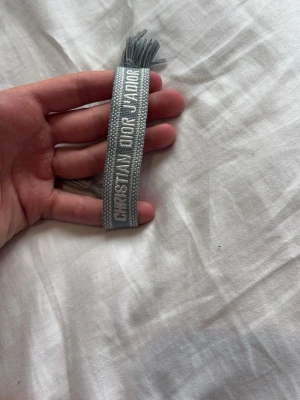 Grått Christian Dior J'Adior armband - Snyggt vävt armband från Christian Dior i ljusgrått med texten 'CHRISTIAN DIOR J'ADIOR' broderad i vitt. Armbandet har dekorativa tofsar i ändarna och är gjort av textilmaterial. Perfekt accessoar för att lyfta din outfit.