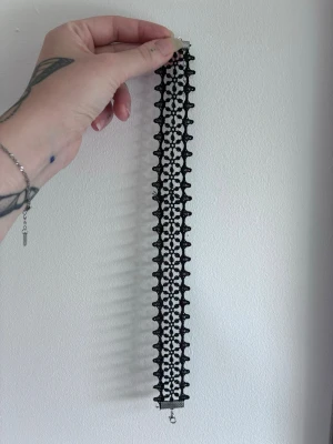 Svart spetschoker halsband - Säljer ett svart choker halsband i bred spets med detaljerat mönster. Halsbandet har metallspänne och justerbar kedja för perfekt passform. Perfekt accessoar för att ge din outfit en edgy och trendig touch.