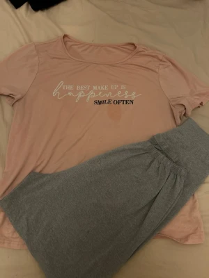 Rosa pyjamas med text och grå byxa - Mysig pyjamas med en ljusrosa t-shirt med trycket 'The best make up is happiness, smile often' och ett par grå mjuka byxor med resår i midjan. Perfekt för chillkvällar hemma. Materialet känns mjukt och bekvämt mot huden.