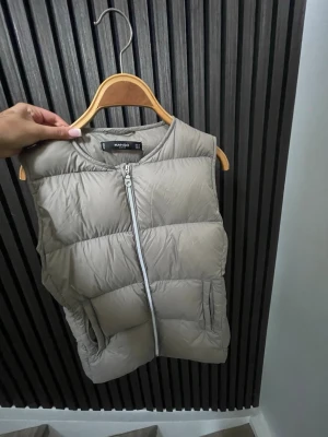 Beige dunväst från Mango - Stilren beige dunväst från Mango med rak passform och breda kanaler. Västen har dragkedja framtill och två fickor på sidorna. Perfekt att slänga över en hoodie eller tröja för extra värme. Enkel och clean design som funkar till det mesta.
