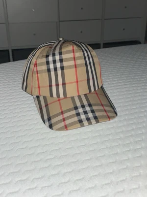 Randig bb keps - Säljer en beige keps med klassiskt rutigt mönster i svart, vitt och rött. Kepsen har böjd skärm och justerbar baksida. Perfekt accessoar för dig som gillar stilrena och tidlösa detaljer. Materialet känns som bomull eller bomullsblandning.