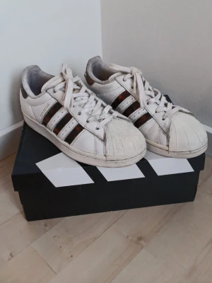 Adidas Superstar vita sneakers - Klassiska Adidas Superstar sneakers i vitt läder med de ikoniska tre ränderna i svart och brunt på sidorna. Skorna har rund tå med gummisnäckskal, vita skosnören och låg profil. Perfekta för en streetwear-look och passar till det mesta😊