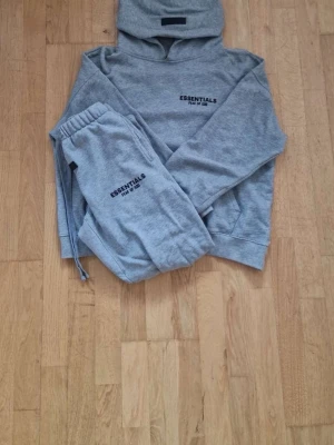 Grått Essentials set med huva - Säljer ett grått set från Essentials Fear of God med hoodie och matchande byxor. Setet har tryckt logga på både tröja och byxa, huva på tröjan och resår i midjan på byxorna. Perfekt för en avslappnad och trendig look.