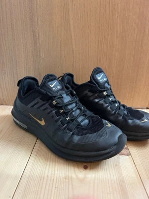 Nike Air Max svarta sneakers - Snygga svarta Nike Air Max sneakers med gulddetaljer och synlig Air-sula. Ovandelen är i mesh och syntetmaterial för bra andningsförmåga och komfort. Klassisk Nike-logga på sidan och tungan. Perfekta för dig som vill ha stilrena och sportiga skor.