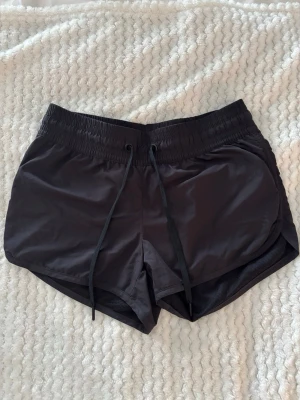 Svarta träningsshorts med snörning - Snygga svarta träningsshorts med elastisk midja och snörning framtill. Shortsen har en sportig look med rundade kanter och meshfoder på insidan för extra komfort. Perfekta för gymmet eller löprundan.