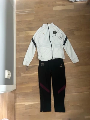 PSG x Jordan träningsset svart/vit - Säljer ett fett PSG x Jordan träningsset med vit zip-jacka och svarta byxor. Jackan har svarta detaljer och PSG-logga på bröstet, byxorna har lila detaljer och Jordan-logga. Perfekt för fotbollsträning eller chill. Materialet är troligtvis polyester. Tröjan har lite fläckar men dom går nog bort i tvätten. Hör av er vid frågor 👍