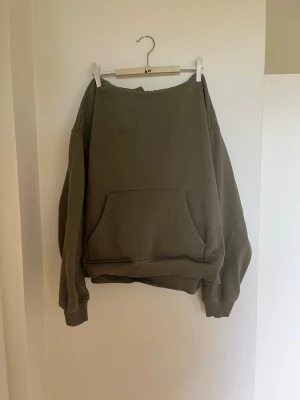 Olivgrön hoodie - Säljer en olivgrön hoodie med en ficka framtill och ribbade muddar vid ärmslut och nederkant. Hoodien har en huva och är tillverkad i mjukt material, perfekt för en avslappnad och chill stil.
