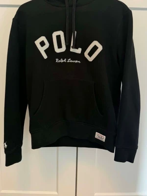 Svart hoodie från Polo Ralph Lauren - Snygg svart hoodie från Polo Ralph Lauren med stor vit POLO-text och broderad logga på bröstet. Klassisk känguruficka framtill, dragsko i huvan och patchdetalj på ärmen. Perfekt för en avslappnad och trendig stil.