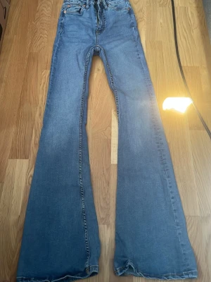 Blå utsvängda jeans med hög midja - Säljer ett par blå jeans med hög midja och riktigt utsvängda ben. Klassisk femficksmodell med knappgylf och kontrastsömmar. Perfekta för dig som gillar retrostil och vill sticka ut lite extra.