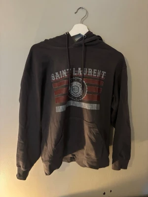 Grå Saint Laurent hoodie med tryck - Snygg grå hoodie från Saint Laurent med huva och dragsnören. Framtill finns ett stort tryck med texten 'SAINT LAURENT' och röda samt vita detaljer. Hoodien har en klassisk känguruficka och är tillverkad i mjukt bomullsmaterial. Perfekt för en avslappnad streetwear-look. Storlek S, skriv vid frågor🔥- vid snabba köp kan priset sänkas🔥