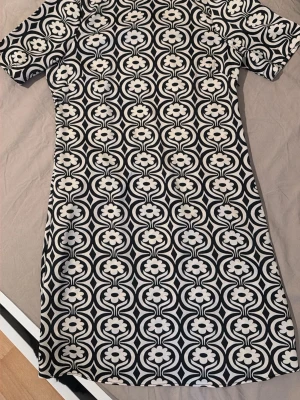 Svartvit mönstrad kortklänning - Kort klänning med svartvitt retroinspirerat blommönster. Klänningen har korta ärmar och en rak passform. Perfekt för dig som gillar grafiska prints och vill sticka ut. Materialet känns mjukt och stretchigt, troligen polyester eller bomull. Köpt från mango 