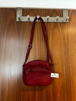 Vinröd axelväska från Glitter - Snygg vinröd axelväska från Glitter i fyrkantig modell med justerbar axelrem. Väskan har en dragkedjeficka framtill och är gjord i ett slätt, konstlädermaterial. Perfekt för att bära det viktigaste och ger en cool touch till din outfit.