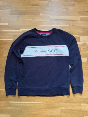 Marinblå sweatshirt från GANT - Snygg marinblå sweatshirt från GANT med vit panel och röda detaljer över bröstet där loggan är tryckt. Tröjan har rund halsringning, långa ärmar och ribbade muddar. Perfekt för en avslappnad och stilren look.