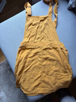 Gul kjol med hängslen - Snygg gul kjol med hängslen och fickor framtill. Den har en avslappnad passform och är gjord i ett mjukt bomullstyg. Perfekt för dig som vill ha en lekfull och trendig look med en twist av retrostil.