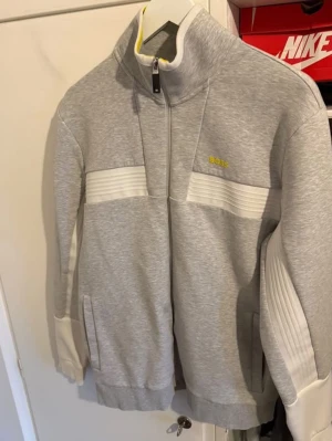 Grå ziptröja från BOSS med vita detaljer - Snygg grå ziptröja från BOSS med vita ribbade detaljer på bröstet och ärmarna. Tröjan har hög krage, dragkedja framtill och fickor på sidorna. BOSS-logga i gult på bröstet och diskret logga på ärmen. Perfekt för en sportig och stilren look. Priset kan diskuteras. Storlek L men passar även M 