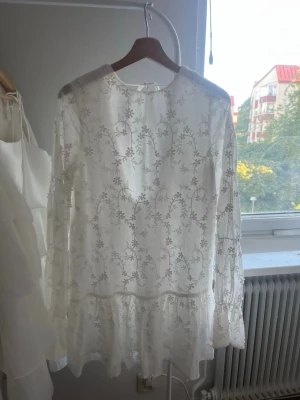 Vit blommig spetsklänning med volang - Romantisk vit kortklänning med blommigt spetsmönster, volangkant nedtill och långärmade ärmar. Klänningen har öppen rygg med knytband i nacken och en luftig, lätt passform som känns perfekt för sommaren. Oanvänd. Nypris: 699kr 