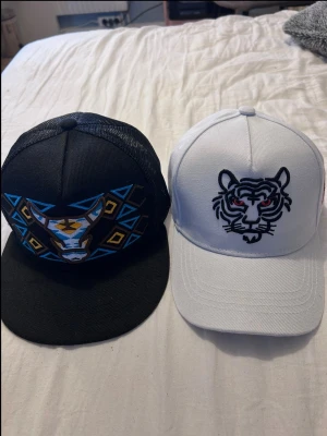 Två kepsar med broderade djurmotiv - Säljer två kepsar: en svart med mesh och broderat tjurmotiv i blått, gult och vitt samt en vit keps med broderad tiger i svart och rött. Båda har böjd skärm och justerbar baksida. Perfekta för dig som gillar unika detaljer och djurmotiv.