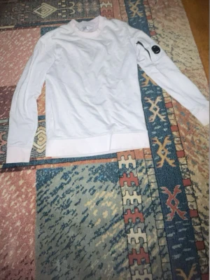 Vit sweatshirt från C.P. Company - Säljer en stilren vit sweatshirt från C.P. Company med deras ikoniska svarta linsdetalj på ärmen. Tröjan har rund halsringning, ribbade muddar och är tillverkad i mjukt bomullsmaterial. Perfekt för dig som gillar minimalistisk och modern streetwear. Storlek xs-s