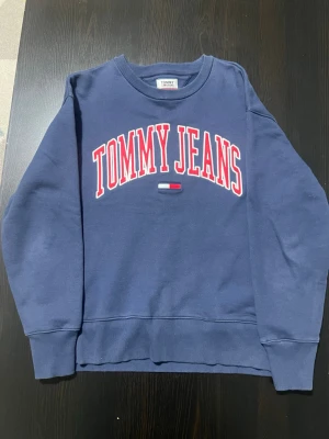 Blå sweatshirt från Tommy Jeans - Mörkblå sweatshirt från Tommy Jeans med stort vitt och rött tryck på bröstet. Klassisk rund halsringning och ribbade muddar vid ärmar och nederkant. Perfekt för en avslappnad och stilren look.