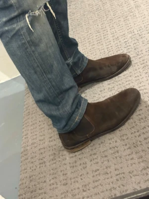 Mörkbruna Chelsea boots i mocka - Säljer ett par stilrena mörkbruna Chelsea boots i mocka med klassisk resår på sidorna och låg klack. Skorna har en smal siluett och passar perfekt till jeans eller chinos. Märket är bytom köpta för 1199 men säljer för endast 349, storlek 43