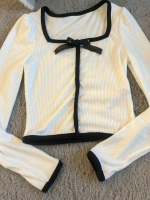 Vit ribbad topp med svart rosett SHEIN - Trendig vit ribbad topp från SHEIN med svarta kanter och en söt svart rosett framtill. Toppen har lång ärm, fyrkantig ringning och croppad passform. Perfekt för dig som gillar detaljer och vill sticka ut lite extra. Köpte den under 2026 men har aldrig kommit till användning.