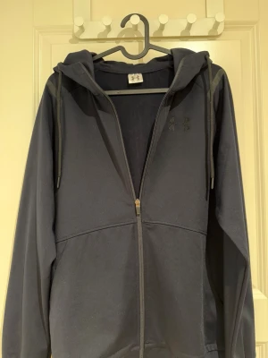 Mörkblå hoodie från Under Armour - Storlek S,  Snygg mörkblå hoodie från Under Armour med dragkedja framtill och justerbar huva med snören. Tillverkad i ett mjukt och bekvämt material, perfekt för chill dagar. Diskret logga på bröstet och två fickor framtill. Klassisk passform och långa ärmar.