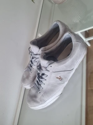 Gråa polo Ralph Lauren skor - Snygga grå sneakers från Polo Ralph Lauren i mockaimitation med vita sulor och klassisk låg profil. Skorna har grå skosnören och en liten metallfärgad logga på sidan. Perfekta för dig som gillar stilrena och tidlösa sneakers.