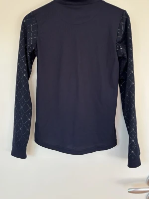 Mörkblå ridströja från Horze - Mörkblå långärmad ridtröja från Horze med hög krage och dragkedja. Ärmarna har ett diskret rutmönster med små symboler, vilket ger en sportig look. Tillverkad i ett stretchigt funktionsmaterial som passar perfekt till träning eller ridning. Andvönd men i gott skick 