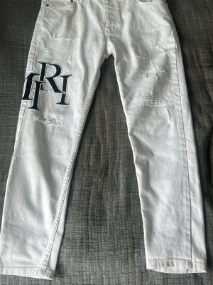 Vita distressed jeans från Amiri - Säljer ett par vita jeans från Amiri med stora svarta bokstavsbroderier på både fram- och baksida. Jeansen har distressed detaljer och raka ben. Perfekta för en trendig streetwear-look.
