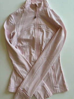 Limited edition Lululemon tröja - Snygg ljusrosa ziptröja från Lululemon med hög krage och figurnära passform. Tröjan har dragkedja framtill, två fickor med dragkedja och tumhål vid ärmsluten. Perfekt för träning eller som ett extra lager. Tillverkad i mjukt, stretchigt material.
