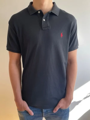 Piké från Polo Ralph Lauren - Säljer dena riktigt fräscha piké från Polo Ralph Lauren. Tröjan är Svart med Röd logga. Den är i storlek M och är i perfekt skick utan defekter! Tveka inte på att höra av dig om du har funderingar!🙌 Priset är ej hugget i sten 🪨 