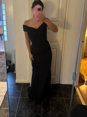 Svart offshoulder långklänning - Elegant svart långklänning med offshoulder-design och figurnära passform. Klänningen har en vacker v-ringad överdel och markerad midja som framhäver siluetten. Perfekt för bal eller festliga tillfällen.