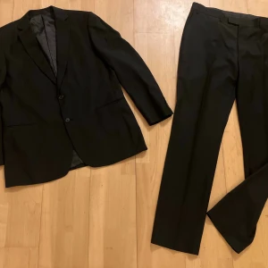 Dressman Svart kostym med kavaj och byxor nyskick!! - Klassisk svart kostym med enkelknäppt kavaj och matchande byxor. Kavajen har två knappar, smala slag och fickor utan lock. Perfekt för dig som vill ha en stilren och tidlös look. Materialet känns slätt och lätt, troligen polyester eller ullblandning. Storleken 54 Regular är en klassisk herrstorlek som motsvarar storlek XL. "Regular" innebär att kavajen har en standardlängd, anpassad för en man som är mellan 175–185 cm lång. Byxor W40 L36 