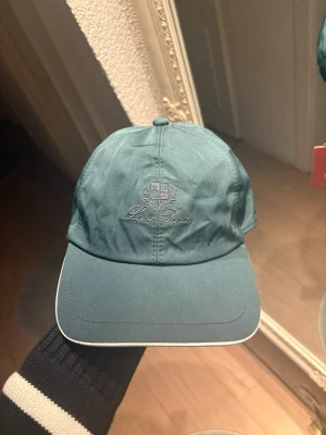 Mörkblå keps från Loro Piana - Snygg mörkblå keps från Loo Piana med broderad logga och text framtill. Skärmen har vit kant och insidan är fodrad med rutigt tyg och orange kant. Justerbar baktill för perfekt passform. Perfekt accessoar för en sportig och stilren look.