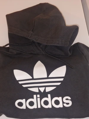 Svart Adidas hoodie - Svart huvtröja med den ikoniska Adidas loggan i vitt tryckt på framsidan. Tillverkad i mjukt bomullsmaterial, perfekt för en avslappnad streetwear-look.