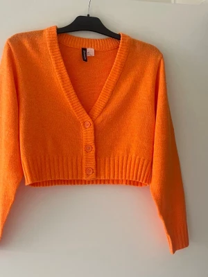 Orange cropped kofta från H&M - Trendig orange cropped kofta från H&M med djup V-ringning och tre knappar framtill. Koftan är stickad i mjukt material och har ribbade muddar vid ärmslut och nederkant. Perfekt för att addera färg till din outfit. 