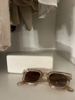  CHIMI 04 - Snygga solglasögon från CHIMI med transparent beige båge och bruna fyrkantiga linser. Ramen är i plast och designen är modern och stilren. Perfekta för dig som vill ha en trendig accessoar som sticker ut.