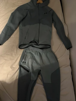 träningsset från Under Armour - Snyggt träningsset från Under Armour med matchande hoodie och byxor. Setet har dragkedja på tröjan, huva och elastisk midja på byxorna. Båda plaggen har Under Armour-logga och byxorna har en praktisk ficka med dragkedja. Perfekt för gym eller löpning.