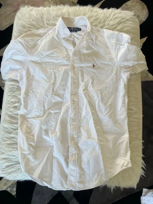 Vit skjorta från Ralph Lauren - Klassisk vit skjorta från Ralph Lauren med button-down krage och broderad logga på bröstet. Skjortan har lång ärm och knappar framtill. Tillverkad i mjuk bomull och har en tidlös, clean look som passar till det mesta.