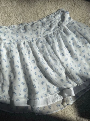 Vit kortkjol med blå blommor - Supersöt vit kortkjol med blått blommönster och volangkant. Kjolen har en bred resår i midjan och är gjord i ett lätt, luftigt material som passar perfekt till varma dagar. Köpt från Gina! Knappt använd💘