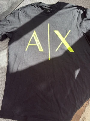 Svart t-shirt från Armani Exchange - Svart t-shirt från Armani Exchange med stort neongult A|X-tryck på bröstet. Slim fit modell med klassisk rund hals och korta ärmar. Tillverkad i mjuk bomull som känns skön mot huden. Perfekt för dig som gillar stilrena och moderna plagg.